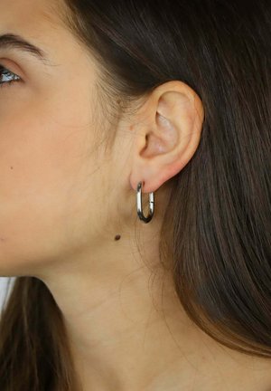 Srebrna metalowa kolczyk kołowy na uchu modelki, charakteryzujący się eleganckim designem, polerowanym wykończeniem i powierzchnią refleksyjną.