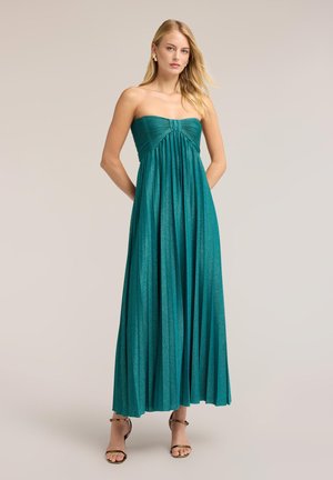 Femme portant une robe longue teal plissée sans bretelles et des sandales dorées à talons hauts, debout devant un fond uni.
