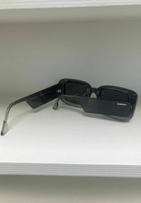 Lunettes de soleil rectangulaires noires avec des branches grises claires et des verres foncés dégradés. Présentent un design géométrique et le nom de la marque "KOMONO" sur le côté.