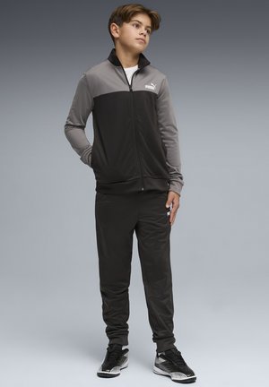 Jongen staat met handen in zijn zakken, gekleed in een zwart-grijs Puma trainingspak en zwarte sneakers, tegen een effen grijze achtergrond.