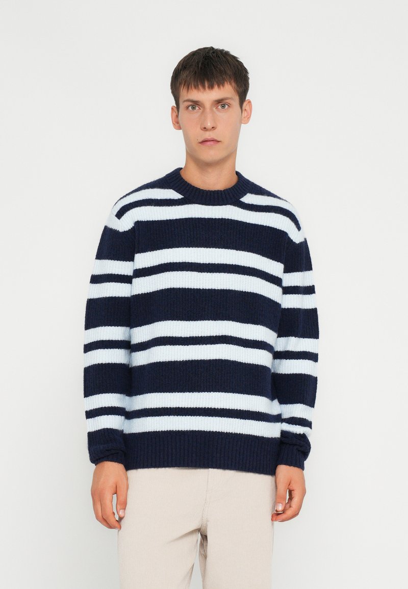 Maglione a righe blu navy e azzurro chiaro, realizzato in maglia, con collo rotondo, polsini e orlo a costine. Il modello indossa pantaloni in corduroy beige.