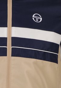 Giubbotto antiviaggio beige e blu navy con strisce orizzontali, finitura testurizzata e logo bianco sulla parte superiore sinistra. Chiusura con zip.