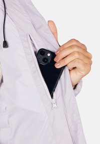 Lys lilla jakke med en synlig lynlåslomme, der holder en sort smartphone. Stoffet ser glat ud med en let skinnende tekstur.