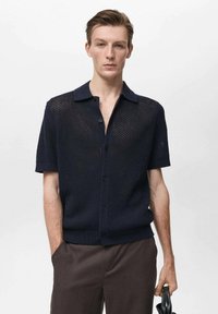 Maglietta in maglia a maniche corte color navy con tessuto a trama aperta, colletto e chiusura a bottoni. Presenta un tessuto testurizzato e una leggera lucentezza. Abbinata a pantaloni scuri.