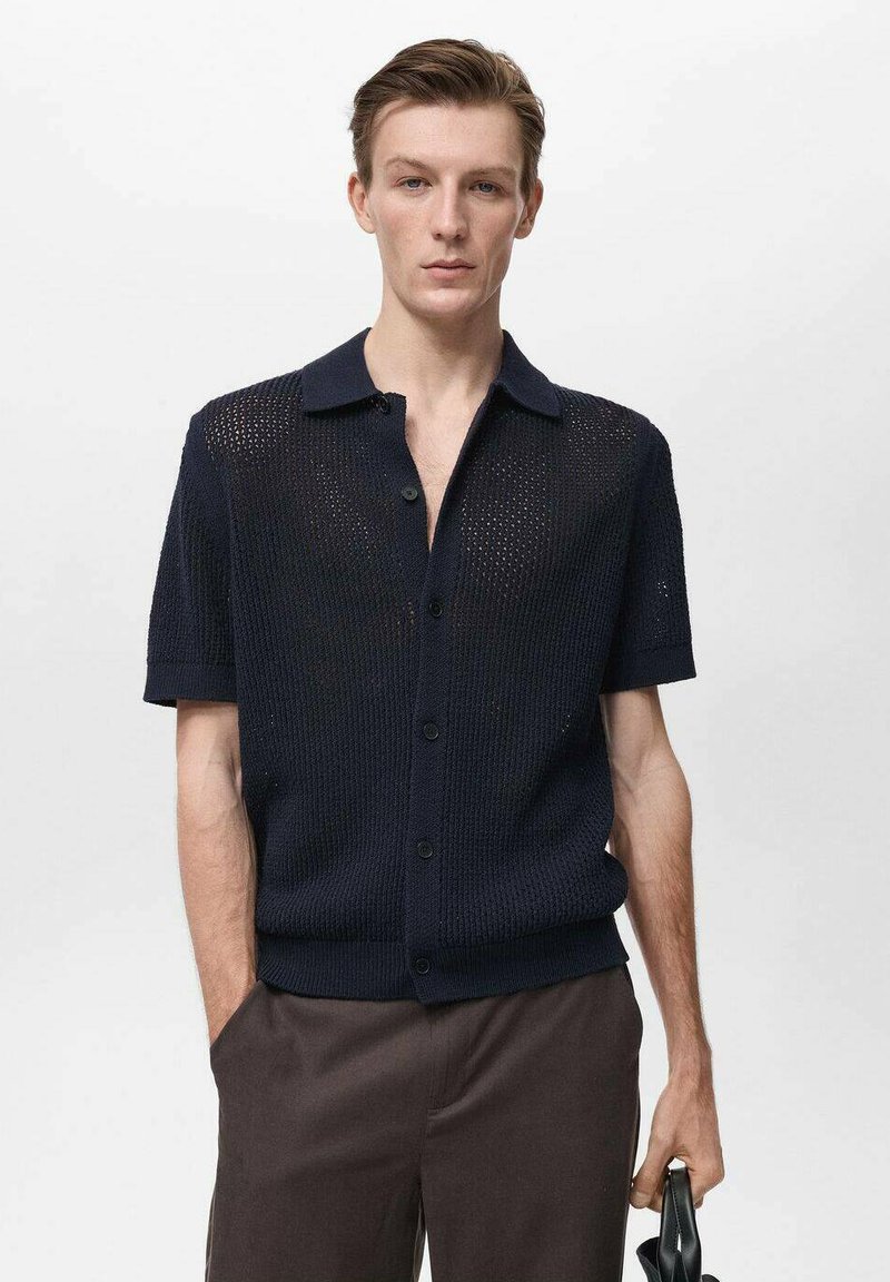 Námořnické tričko s krátkým rukávem z pleteniny s otevřenou tkaninou, límcem a button-down zapínáním. Má texturovanou látku a jemný lesk. Stylizováno s tmavými kalhotami.
