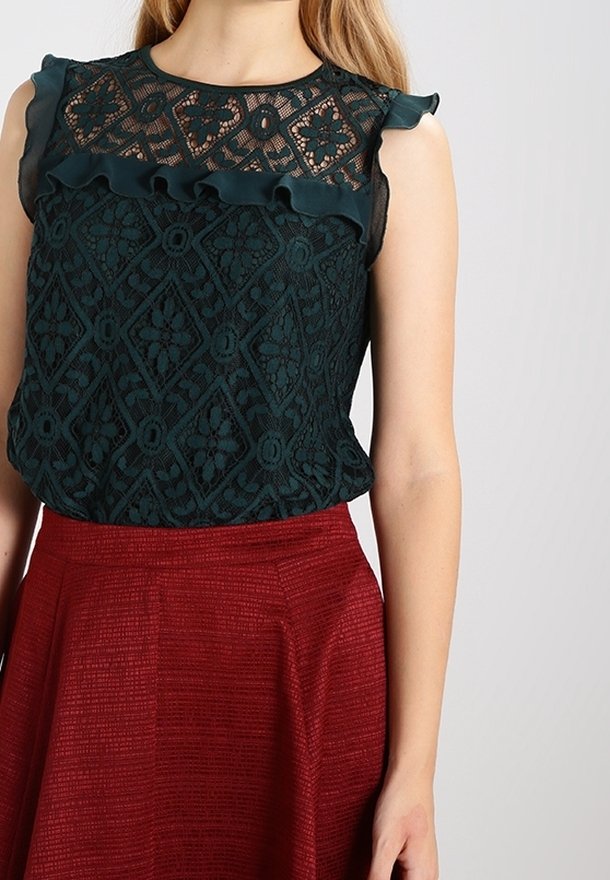 Top de encaje verde oscuro con detalles de volantes en los hombros, combinado con una falda roja texturizada. El top presenta un patrón de diamantes.