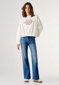 Sudadera blanca con el logo rojo de "Pepe Jeans London", combinada con vaqueros azules de campana. Los vaqueros tienen un dobladillo ligeramente deshilachado y una cintura ajustada.