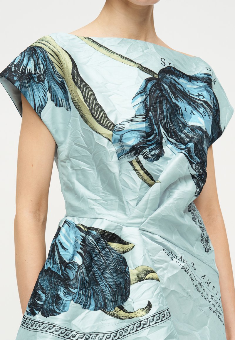 Hellblaues Kleid mit großen blauen und schwarzen Blumenmustern sowie schwarzem Schriftzug, getragen von einer Person mit sichtbaren nackten Armen.