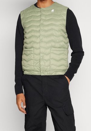 Uomo che indossa un gilet trapuntato verde chiaro sopra una maglia nera a maniche lunghe e pantaloni cargo neri, con una mano in tasca.