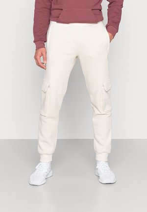 Homme portant un pantalon jogger cargo crème avec des poches latérales, un sweat bordeaux et des baskets blanches sur un fond clair uni.