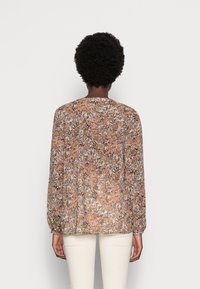 Femme aux cheveux courts et bouclés portant un blouse à manches longues à fleurs dans des tons de rose et de marron, accompagnée d'un pantalon blanc, se tenant de dos.