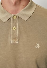 Béžová polo košile vyrobená z texturované látky, s límečkem, dvěma žlutými knoflíky a malým bílým logem na hrudi.