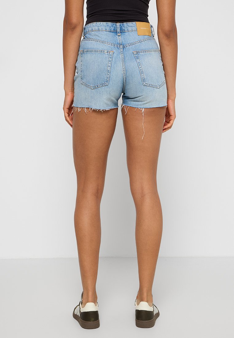 Pantalones cortos de mezclilla azul claro con dobladillos deshilachados, bolsillos traseros y una etiqueta de cuero marrón en la cinturilla. Combinados con zapatillas blancas y oliva.