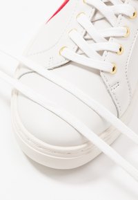 Baskets blanches avec lacets blancs passés dans des œillets dorés, gros plan sur les détails de couture à l’avant et sur le côté.
