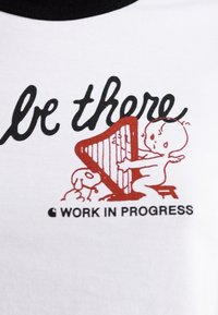 Witte T-shirt met zwarte halslijn, met een rode illustratie van een baby die harp speelt, vergezeld door een hond, en de tekst "WORK IN PROGRESS."