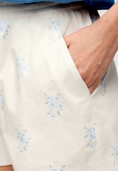 Main insérée dans la poche d'un short en tissu blanc avec un motif floral bleu clair et une taille élastique.
