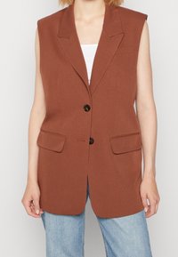 Brun ärmlös blazer med två framfickor, slagkrage och två svarta knappar; kombinerad med en vit topp och blå jeans.