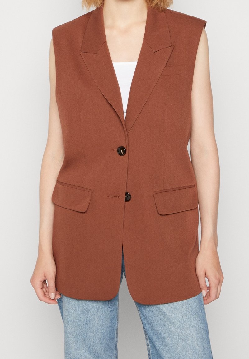 Brun ärmlös blazer med två framfickor, slagkrage och två svarta knappar; kombinerad med en vit topp och blå jeans.