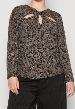Femme portant un haut à manches longues marron et noir à motifs avec des découpes près du décolleté et un pantalon texturé noir.