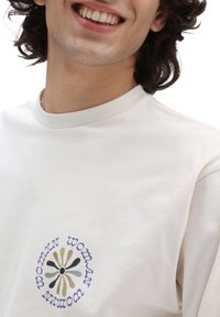 Camisa de manga larga color crema con cuello redondo y un gráfico de flores que presenta el texto "mujer" y "womxn" en tonos negros, morados y verdes.
