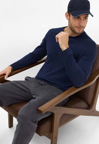 Marineblauwe longsleeve shirt met een klein zakje, gecombineerd met donkergrijze broek. Man zit op een bruin gestoffeerde stoel met houten armleuningen.