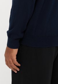 Maglione a maglia blu navy con vestibilità comoda e polsini a coste, abbinato a pantaloni neri. Primo piano sul dettaglio della manica e della mano.