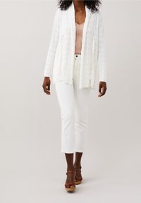 Cardigan bianco a maglia con apertura frontale, motivo testurizzato, indossato su una maglietta rosa chiaro. Abbinato a pantaloni bianchi tagliati e sandali marroni.