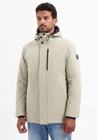 Beige waterdichte jas met een capuchon, een ritszak op de borst en knoopsluitingen. Heeft een slank ontwerp en een zachte textuur met zwarte accenten.