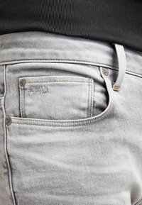 Vaqueros de mezclilla gris claro con un pequeño bolsillo trasero, remaches metálicos y una textura sutilmente desgastada. Presillas para cinturón en la cintura que mejoran la funcionalidad del diseño.