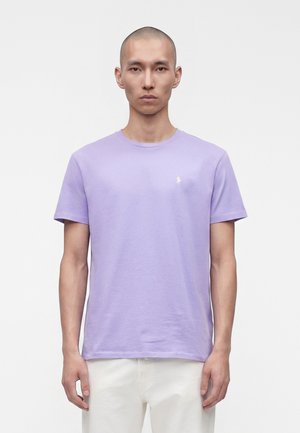 Mann mit rasiertem Kopf, der ein schlichtes lavendelfarbenes T-Shirt mit Rundhalsausschnitt und weiße Hose trägt, steht vor einem weißen Hintergrund.