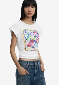 Weißes T-Shirt mit kurzen Ärmeln, das mit einem bunten, abstrakten Grafikdruck mit Herzen und einem Randdesign verziert ist. Kombiniert mit hochgeschnittenen blauen Jeans.