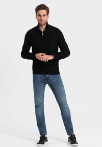 Maglione nero in maglia con collo metà zip, indossato sopra jeans azzurri chiari. Il modello indossa anche scarpe da ginnastica nere e si trova in una posa neutra.