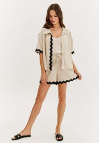 Krémová krémová směs krátkého setu se černým vlnkovaným lemem, košilí s krátkým rukávem na knoflíky, šortkami s elastickým pasem a plochými černými sandály.