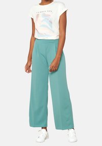Mujer usando pantalones anchos de color verde azulado, zapatillas blancas y una camiseta blanca con un diseño abstracto en tonos pastel y el texto "La Dolce Vita."