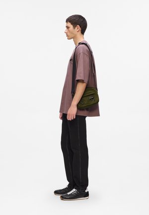 Joven con una camiseta oversized malva, pantalones negros y zapatillas de deporte, lleva una bolsa cruzada de color verde oliva frente a un fondo blanco liso.