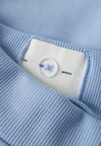 Tela de color azul claro con una textura acanalada. Presenta una etiqueta blanca con un botón circular transparente y detalles cosidos.