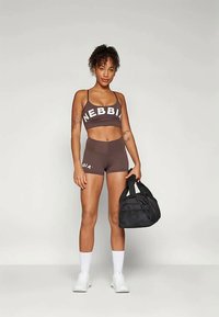 Femeie îmbrăcată într-un top sport maro și pantaloni scurți, ținând o geantă de sală neagră, stând împotriva unui fundal simplu deschis, purtând șosete albe și adidași.