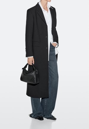 Person iført en lang sort frakke, hvid skjorte med knapper, vidde bukser i mørk denim, sorte spidse sko, der holder en fletsort taske.