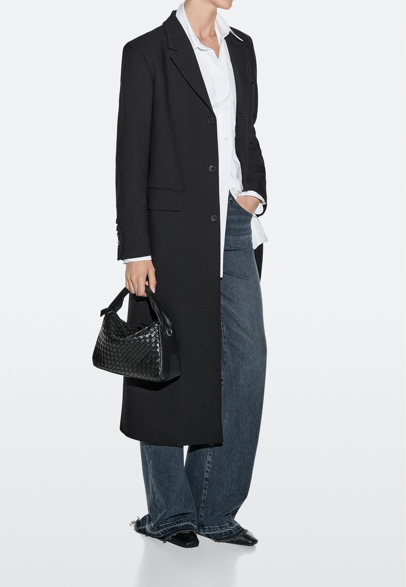 Person trägt einen langen schwarzen Mantel, ein weißes Hemd mit Knöpfen, weite dunkle Jeans, schwarze spitze Schuhe und hält eine schwarze, geflochtene Handtasche.