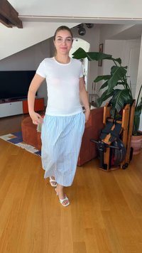 Γυναίκα με λευκή μπλούζα και μπλε ριγέ φούστα περπατά σε ξύλινο πάτωμα σε σαλόνι με φυτά και εξοπλισμό γυμναστικής.
