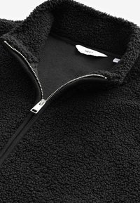 Juoda vilnos zip-up striukė su aukštu apykakle, tekstūruotu paviršiumi, sidabriniu užtrauktuku ir vidinė linija su lygiu paviršiumi.