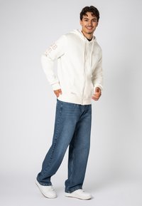 Weißer Reißverschluss-Hoodie mit Kapuze und Kordelzug, versehen mit bedrucktem Text auf den Ärmeln, kombiniert mit lockeren blauen Jeans und weißen Sneakers.