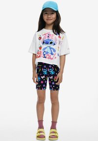 Camiseta blanca de manga corta con un gráfico colorido de Stitch, combinada con pantalones cortos azules marinos de longitud a la rodilla con patrones florales. Calcetines rosas y sandalias amarillas.