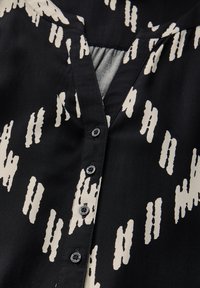 Blusa nera con motivo astratto bianco, tessuto leggero, scollo a V, sei bottoni neri sul davanti e texture liscia.