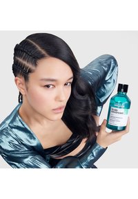 Teal schampo-flaska med svart lock märkt "Scalp Advanced" som hålls i en persons hand, klädd i en glänsande blå topp med texturerade ärmar.