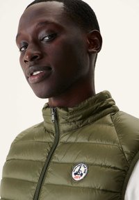 Olijfgroene, gewatteerde vest met hoge kraag, ritssluiting aan de voorkant en logo-patch. Textiel met structuur, lichte ontwerp dat geschikt is voor laagjes.