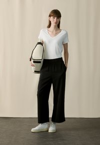 Weißes V-Ausschnitt-T-Shirt, schwarze locker sitzende Hose, weiße Sneakers mit strukturierten Sohlen und cremefarbener Schultertasche mit schwarzen Akzenten.