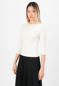Mulher com cabelo loiro liso vestindo uma blusa justa de manga longa creme e uma saia plissada preta de cintura alta, em pé contra um fundo branco.