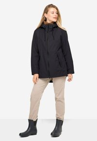 Chaqueta negra impermeable con capucha, cierre frontal y bolsillos laterales, combinada con pantalones beiges y botas de goma negras. Textura suave.