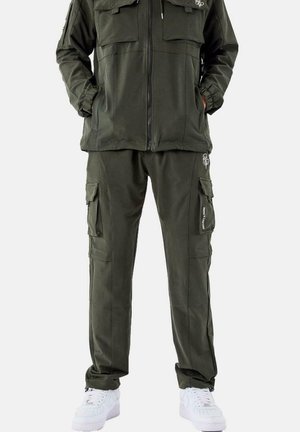 Ensemble de veste et pantalon cargo vert olive en tissu doux. La veste possède deux poches poitrine ; le pantalon est doté de plusieurs poches latérales, associé à des baskets blanches.